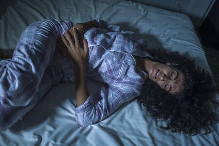 dramatic night lifestyle portrait of young sad and depressed latin woman with curly hair sleepless in bed suffering excruciating period pain holding her belly in fetal positionの写真素材