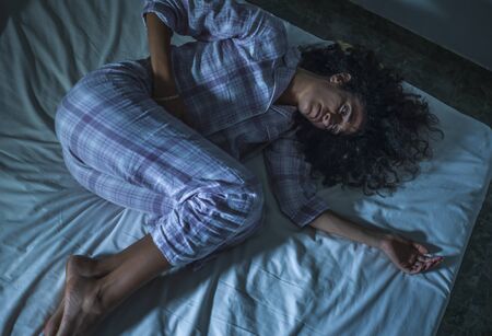 dramatic night lifestyle portrait of young sad and depressed middle eastern woman with curly hair sleepless in bed suffering excruciating period pain holding her belly in fetal positionの写真素材