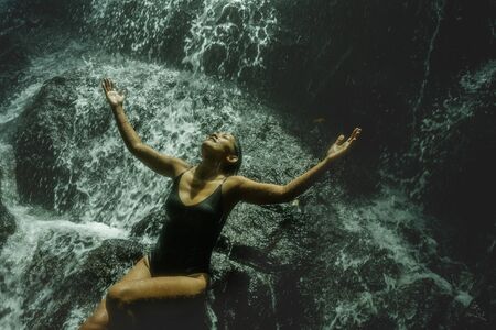 young attractive and blissful Asian Indonesian woman overwhelmed by the beauty of natural waterfall...