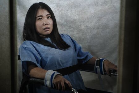 young crazy and mentally insane Asian woman restrained in wheelchair at mental hospital suffering psychiatric disorder as schizophrenia looking catatonic in Korean horror movie styleの写真素材