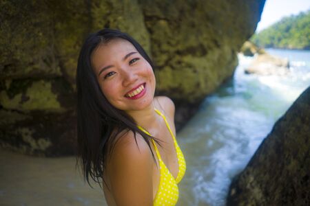 outdoors portrait of young happy and beautiful Asian Chinese woman in yellow bikini excited having holidays at paradise beach island playing in the sea enjoying idyllic travel destinationの写真素材