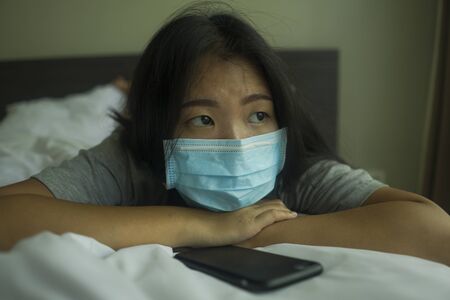 pandemic and virus outbreak - young beautiful scared and worried Asian Korean woman in medical mask checking online news with mobile phone on bed lockdown at home in quarantineの写真素材