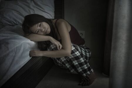 dramatic dark portrait of young attractive depressed and sad Asian Chinese woman on bedroom floor feeling worried and scared suffering depression problem and anxiety leaning on bedの写真素材