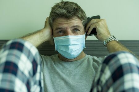 virus outbreak - dramatic portrait of sick man in medical facial mask sitting on bed scared and...