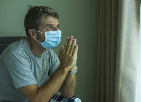 virus outbreak - dramatic portrait of sick man wearing medical facial mask sitting on bed scared and...