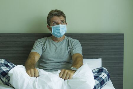 virus outbreak - dramatic portrait of sick man wearing medical facial mask sitting on bed scared and worried infected by coronavirus suffering lockdown alone at home put in quarantineの写真素材