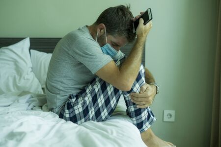 virus outbreak - dramatic portrait of sick man in medical facial mask sitting on bed scared and worried infected by coronavirus suffering lockdown alone at home in quarantineの写真素材