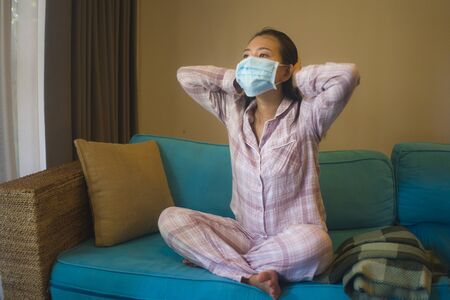quarantine and home lockdown - young beautiful stressed and worried Asian   woman in pajamas on couch in surgical mask feeling sad and scared about covid-19 virus outbreakの写真素材
