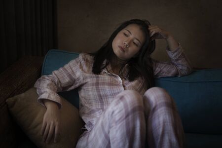 dramatic dark portrait of young beautiful sad and depressed Asian   woman in pajamas at home sitting by living room couch feeling unhappy and confused suffering broken heart painの写真素材