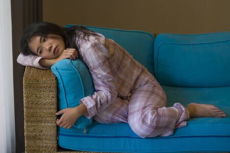 indoors lifestyle portrait of young beautiful sad and depressed Asian Chinese woman in pajamas at home sitting by living room couch feeling unhappy and confused suffering broken heart painの写真素材