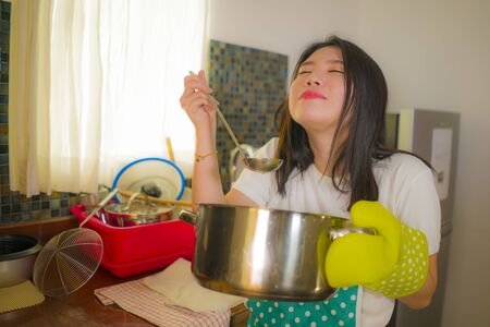 Beautiful domestic chef at work - lifestyle portrait of young pretty and happy Asian Chinese woman in apron holding cooking pot tasting delicious soup smiling satisfied as home cook housewifeの写真素材
