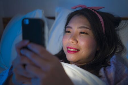 lifestyle portrait of young beautiful happy and relaxed Asian Chinese woman on bed playing with mobile phone on bed in the dark in internet and social media addiction and overuseの写真素材
