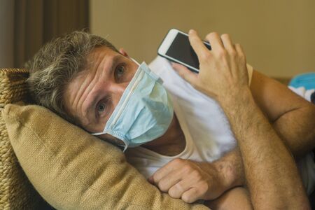 following online news on covid-19 virus pandemic - man with face mask in home quarantine lockdown checking internet information on the contagious disease scared and worriedの写真素材