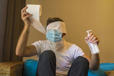 toilet paper sold out paranoia during covid-19 quarantine - crazy man in protective mask in home lockdown with head wrapped in tissue paper roll during coronavirus pandemic outbreakの写真素材