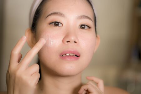 home lifestyle portrait of young happy and beautiful Asian Korean woman at bathroom mirror applying skin care moisturizer and anti-aging product in beauty and skincare conceptの写真素材