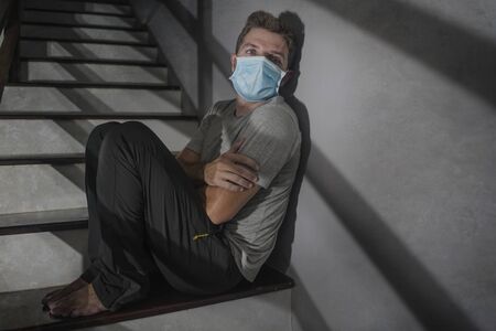 dark portrait of young scared and worried man in protective mask sitting on stairs at home staircase during lockdown and quarantine for covid-19 virus pandemic in dramatic lightの写真素材