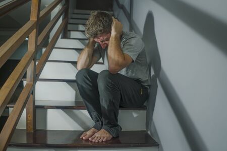 horror movie style portrait of sad and desperate man suffering depression problem or mental disorder sitting on staircase at home hopeless crying overwhelmed and helplessの写真素材