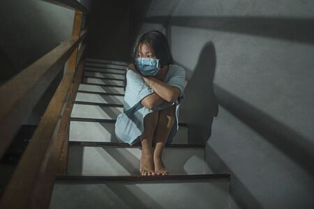 sad portrait of young scared and worried Asian Korean woman in protective mask sitting on stairs at...