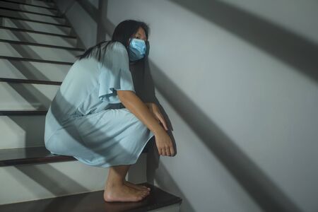 sad portrait of young scared and worried Asian  woman in protective mask sitting on stairs at home lockdown and quarantine during covid-19 virus pandemic in dramatic lightの写真素材