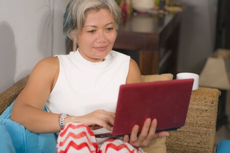 attractive and happy middle aged woman on her 50s working on laptop computer at living room couch...