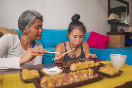 Asian cheereful female friends having Japanese sushi for lunch - happy and attractive woman and her girlfriend eating sushi rolls at home enjoying together healthy delicious food on couchの写真素材