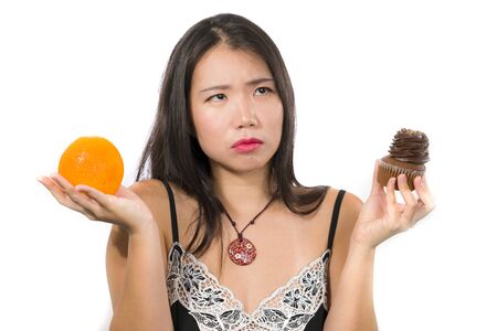 young attractive and happy Asian Chinese woman holding delicious and tempting chocolate cupcake full of calories and fresh healthy orange fruit in low calories versus sugary junk foodの写真素材