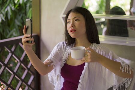 outdoors holiday lifestyle portrait of young beautiful and happy Asian Chinese woman having morning coffee taking selfie with mobile phone for social media at home terrace or hotel room balconyの写真素材