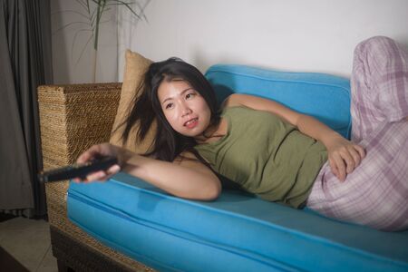 home lifestyle portrait of young happy and beautiful Asian Chinese woman watching TV  at living room...