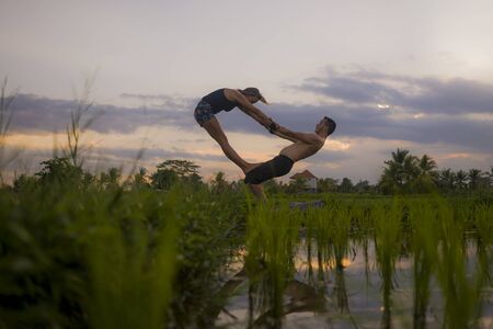 outdoors sunset acroyoga workout silhouette- young happy and fit couple practicing acro yoga drill at beautiful rice field enjoying nature and healthy lifestyle doing acrobatic poseの写真素材