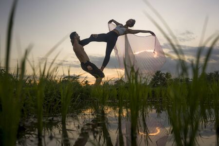 outdoors sunset acroyoga workout - young happy and fit couple practicing acro yoga drill at beautiful rice field enjoying nature doing acrobatic pose with woman in wings in freedom conceptの写真素材