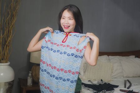 home lifestyle portrait of young happy and sweet Asian Japanese woman trying different clothes and new dress looking in bedroom mirror deciding what to wear for her dateの写真素材