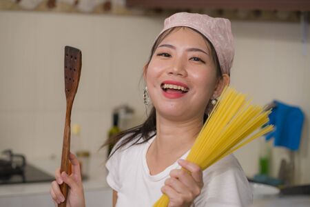 beautiful and happy Asian woman preparing noodles - domestic lifestyle portrait of young sweet and playful Korean girl at home kitchen cooking pasta smiling playful holding spaghettiの写真素材