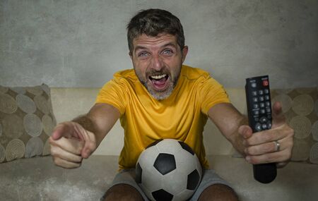 young excited and crazy happy football supporter man watching soccer game on television at living room sofa holding TV remote celebrating team scoring goal feeling intense emotionの写真素材