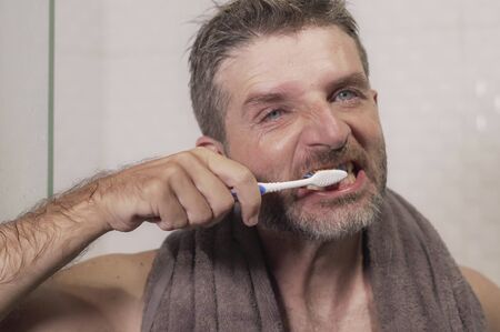 home lifestyle portrait of young attractive and happy man with towel on his neck brushing his teeth in the bathroom relaxed and cheerful enjoying dental care routine の写真素材