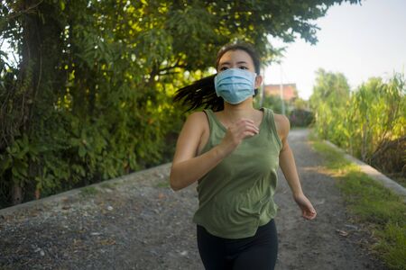 Asian woman in face mask running workout during covid-19 deconfinement- young dedicated Korean girl jogging at beautiful city park on sunset enjoying fitness in anti pollution facial maskの写真素材