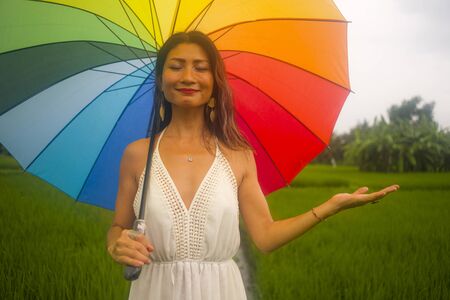 outdoors holidays portrait of young happy and beautiful Asian Korean woman in white dress and rainbow umbrella enjoying nature carefree at tropical green field walking around の写真素材