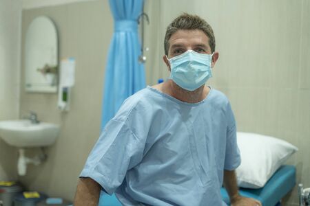 attractive and scared man infected by covid-19 - dramatic portrait of adult male in face mask receiving treatment at hospital suffering respiratory disease sitting on bed worried の写真素材