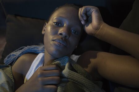 Millennial generation Afro American girl at night suffering depression - young attractive sad and depressed black woman lying thoughtful and worried on sofa couch in pain and fearの写真素材
