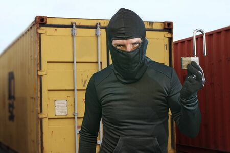 thief at work - criminal man in black covered with balaclava mask holding unlocked padlock at...