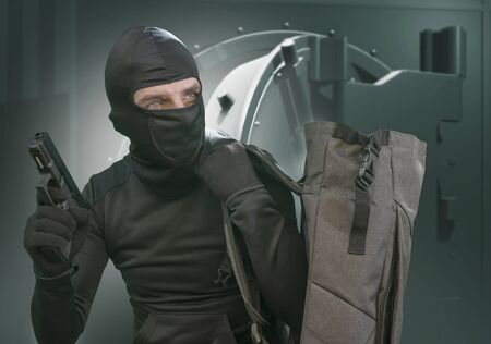 heist and robbery - portrait of man in balaclava mask holding gun in front of security metal vault...