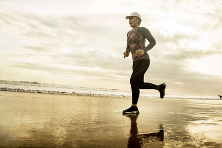 Silhouette of middle aged woman running on the beach - 40s or 50s attractive mature lady doing jogging workout enjoying fitness and healthy lifestyle at beautiful sea sunset landscapeの写真素材