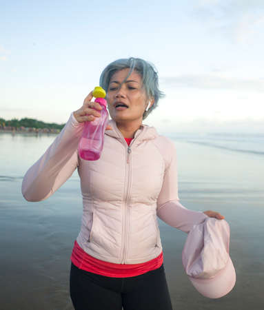 happy tired and thirsty middle aged woman drinking water after beach running workout - 40s or 50s attractive mature lady with grey hair re hydrating  exhausted after joggingの写真素材