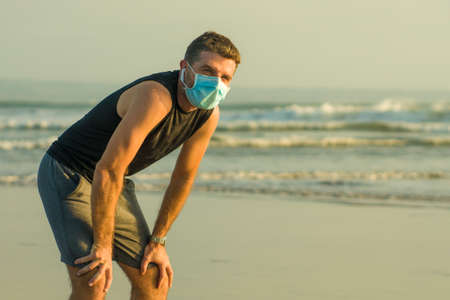 New normal beach jogging workout - young fit and attractive man in face mask breathing tired doing cool off after running on sea training again after covid19 lockdownの写真素材