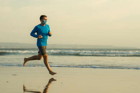 New normal beach jogging workout - young fit and attractive man in face mask running barefoot on sea training after covid19 lockdown feeling free outdoors in healthy lifestyle conceptの写真素材