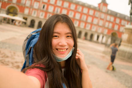 new normal holidays travel in Europe - young happy and beautiful Asian Chinese tourist woman wearing mask taking selfie with mobile phone enjoying city sightseeing and urban tourismの写真素材