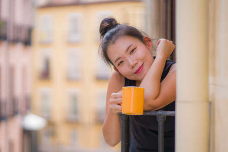 young beautiful and happy Asian Chinese woman enjoying city view from hotel room balcony in Spain during holidays trip in Europe drinking coffee relaxed in urban backgroundの写真素材