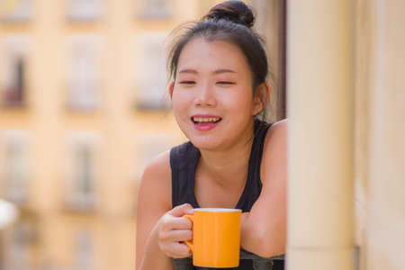young beautiful and happy Asian Chinese woman enjoying city view from hotel room balcony in Spain during holidays trip in Europe drinking coffee relaxed in urban backgroundの写真素材