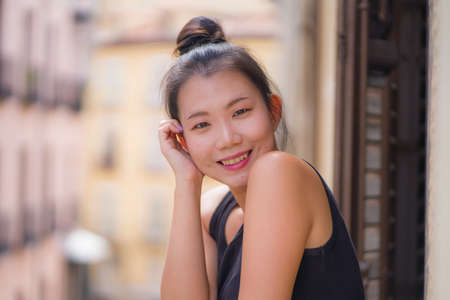 young happy and beautiful Asian Korean woman in hair bun enjoying city view from hotel room balcony in Spain during holidays trip in Europe smiling cheerful in urban backgroundの写真素材