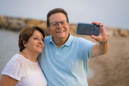 happy pensioner woman and her husband taking romantic walk taking selfie - happy retired mature couple walking on the beach during holidays taking self portrait with mobile phoneの写真素材