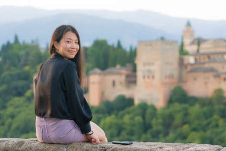 lifestyle portrait of young beautiful and happy Asian Japanese tourist woman during Summer holidays in Europe sitting cheerful enjoying beautiful view of Alhambra in Granada Spainの写真素材
